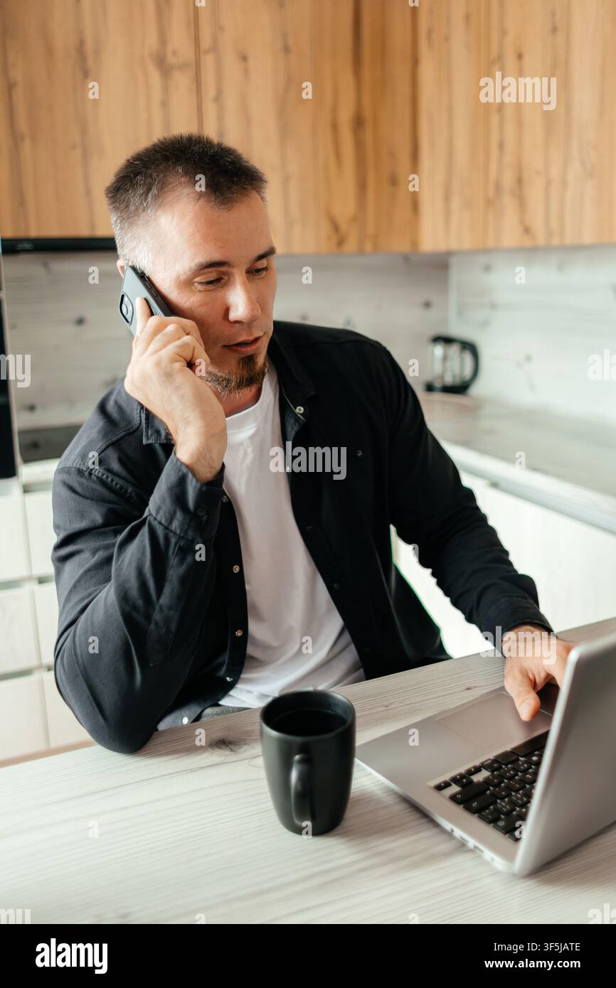 Mann, der von zu Hause aus arbeitet, Multitasking mit einem Laptop und einem Telefonat. Er sitzt an einem Holztisch und konzentriert sich auf seine Aufgaben, was die Dynamik der Fernarbeit veranschaulicht Stockfoto
