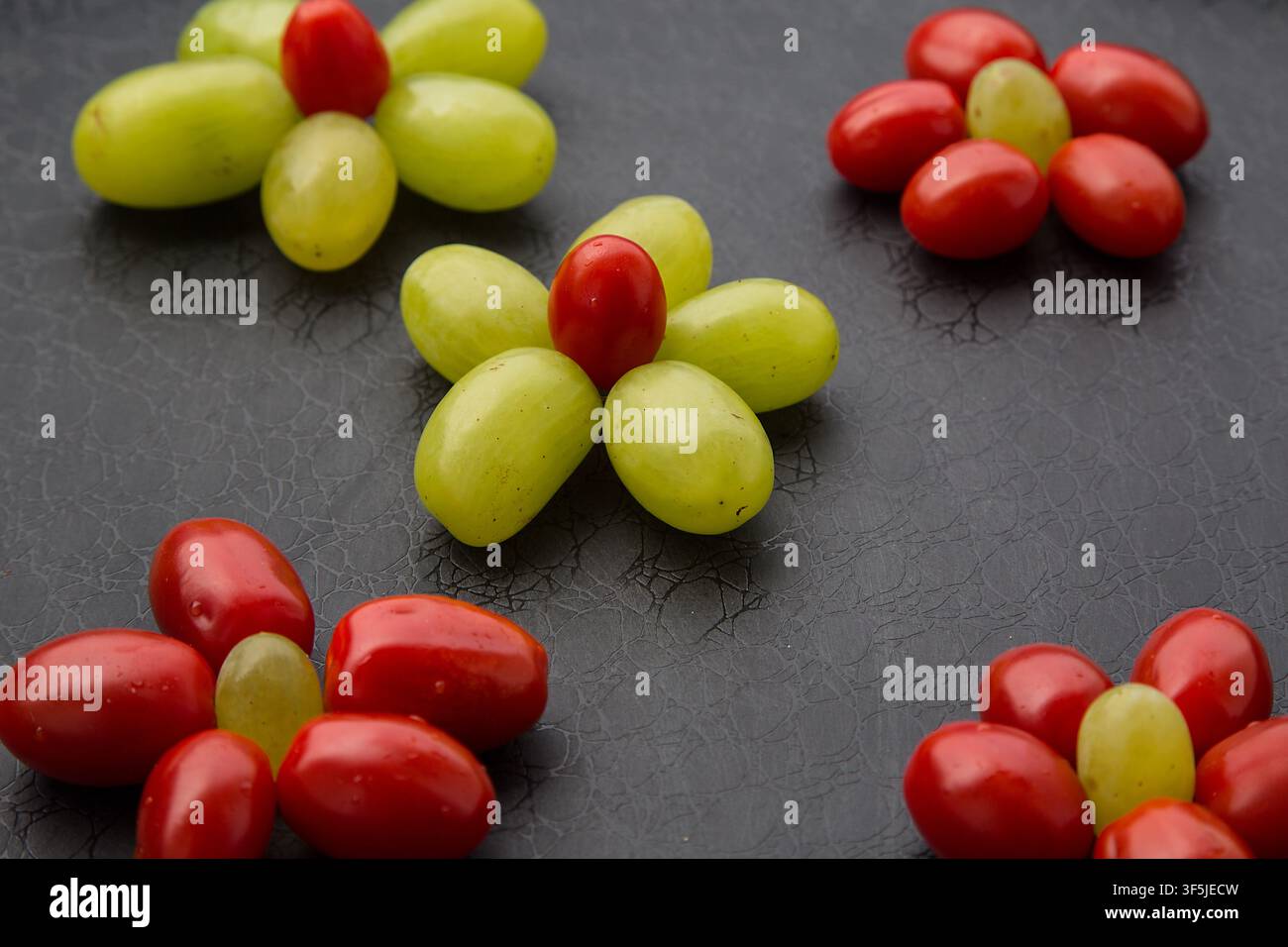 Kreative Anordnung von grünen Trauben und roten Cocktailtomaten, die wie bunte Blumen geformt sind, auf einem schwarzen strukturierten Hintergrund. Dieses verspielte gesunde Essen com Stockfoto