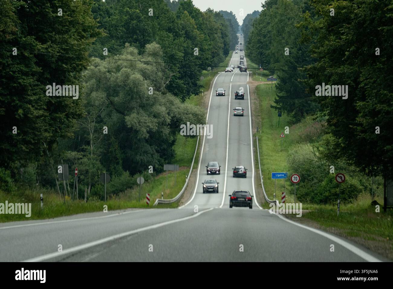 Lettland – 1. August 2026: Autos fahren eine hügelige zweispurige Autobahn durch eine dicht bewaldete ländliche Landschaft. Stockfoto