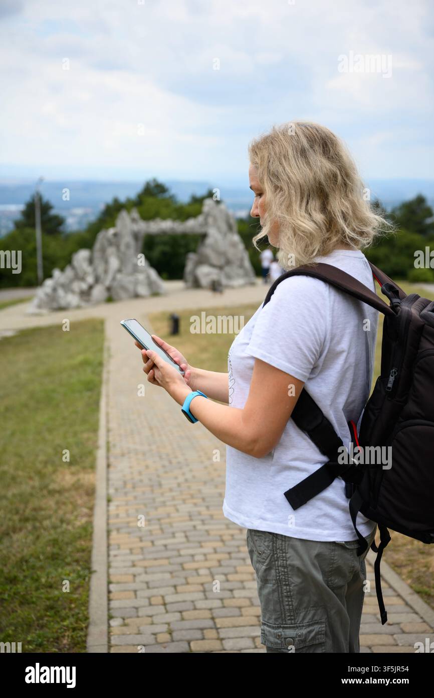 Eine vertikale Fotografie einer mittelalten kaukasischen Frau, die auf einem gepflasterten Außenweg steht und auf ihr Smartphone schaut. Sie trägt ein weißes T-s Stockfoto