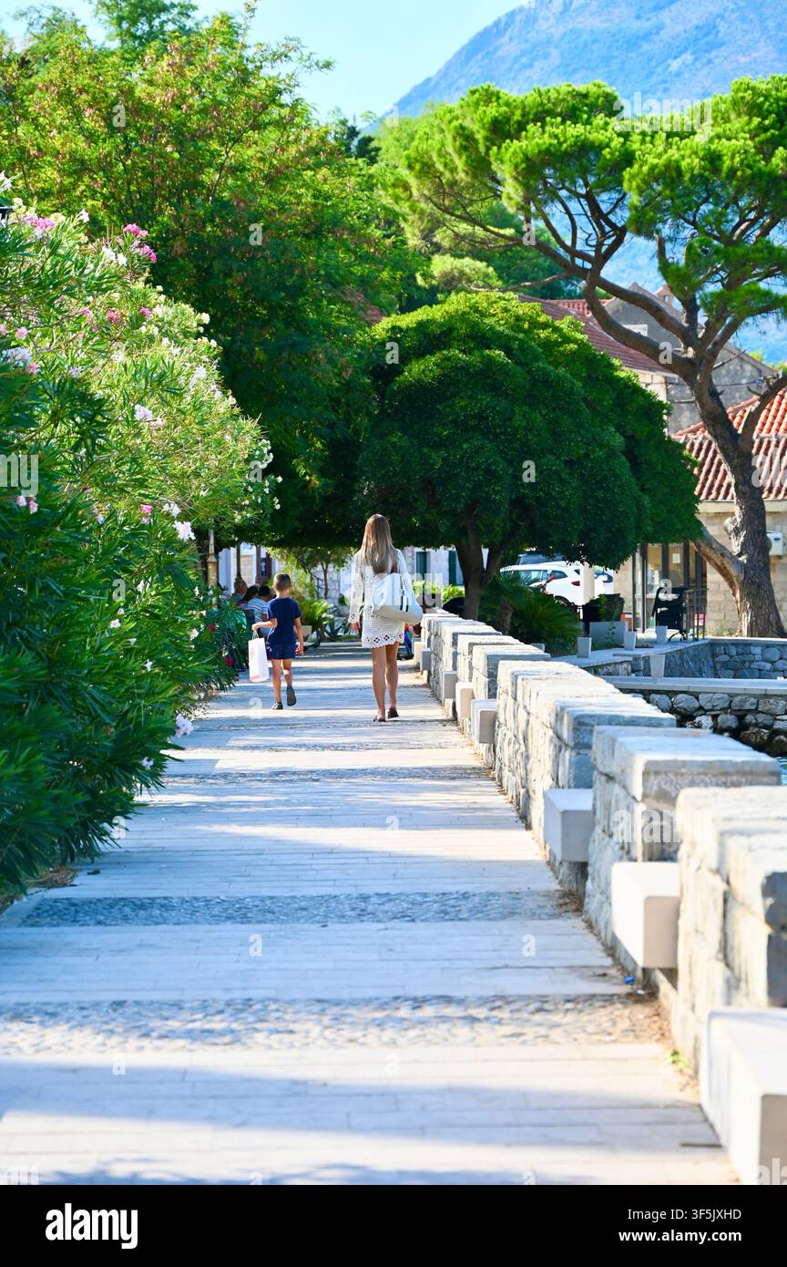 Eine vertikale Tagesaussicht von Menschen, die entlang eines malerischen Steinweges und einer Promenade mit niedrigen Steinmauern, üppigen grünen Bäumen und Büschen weggehen, wobei im oberen Hintergrund Berghänge sichtbar sind. Stockfoto
