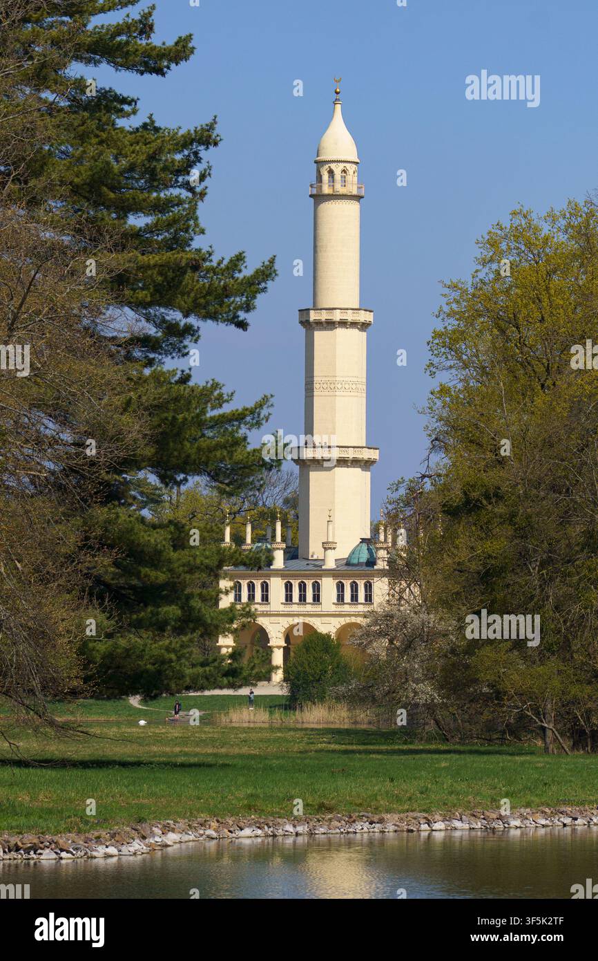 Eine vertikale Augenhöhe-Landschaftsfotografie des historischen maurischen Lednice-Minarettturms, eingerahmt von hohen grünen Bäumen über einem ruhigen Teich auf dem c Stockfoto