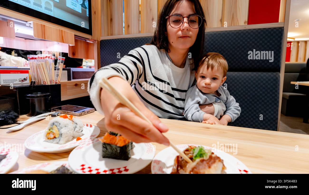 Eine Familie genießt das Abendessen in einem Sushi-Restaurant mit Fließband in Tokio, Japan. Die Mutter genießt frisches Sushi, während sie ihr Kind hält, und fängt ein mich Stockfoto