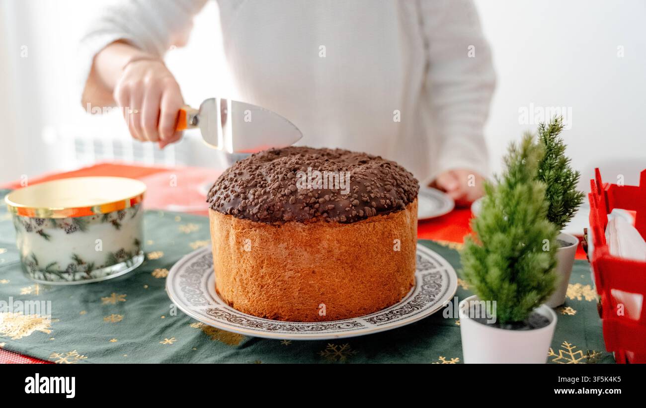 Eine Hand schneidet einen frischen Panettone-Kuchen auf einem geschmückten Esstisch, der für ein Neujahrsfrühstück zu Hause vorbereitet ist, mit festlichen Dekorationen, Kerze und gemütlichem i Stockfoto