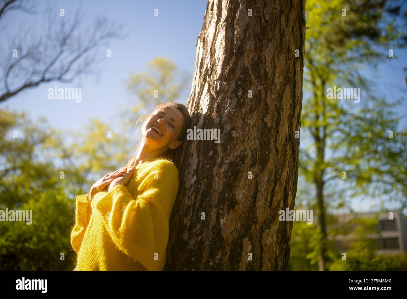 Eine strahlende Frau in einem gelben Pullover, die an einem Baum lehnt, mit geschlossenen Augen lächelt und die reine Lebensfreude, geistige Gesundheit und Harmonie einfängt Stockfoto