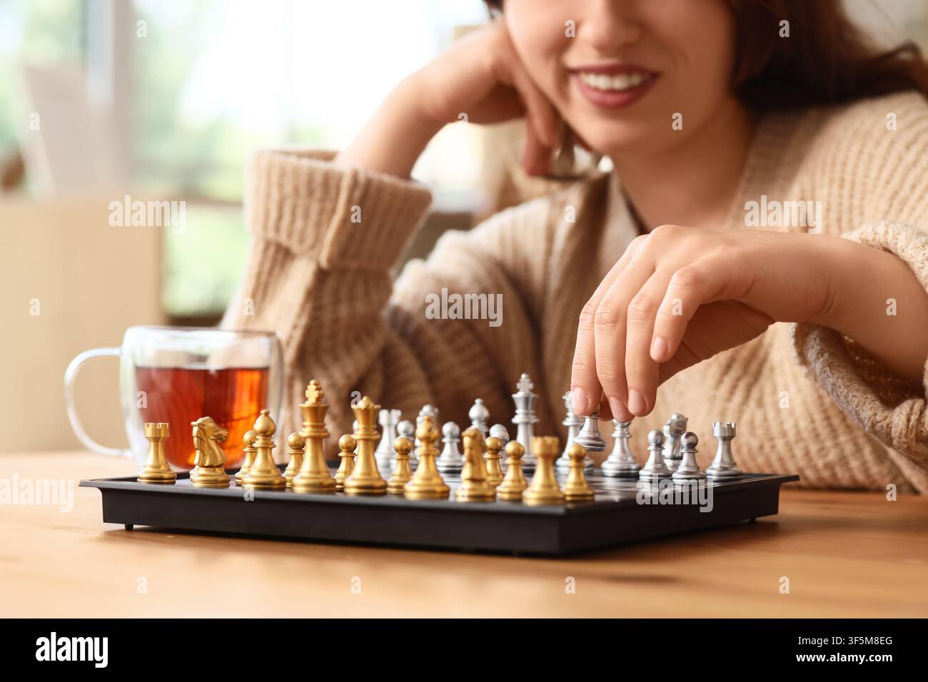 Lächelnde Frau spielt Schach mit einer Tasse Tee, Strategie und intellektuelle Herausforderung als Konzept, Freizeitaktivität zu Hause Stockfoto