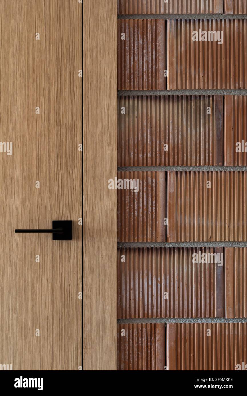 Nahaufnahme einer Holztür mit mattschwarzem Griff neben einer strukturierten braunen Ziegelwand mit vertikalem Rillenmuster und warmen natürlichen Tönen, kombiniert mit glattem Stockfoto