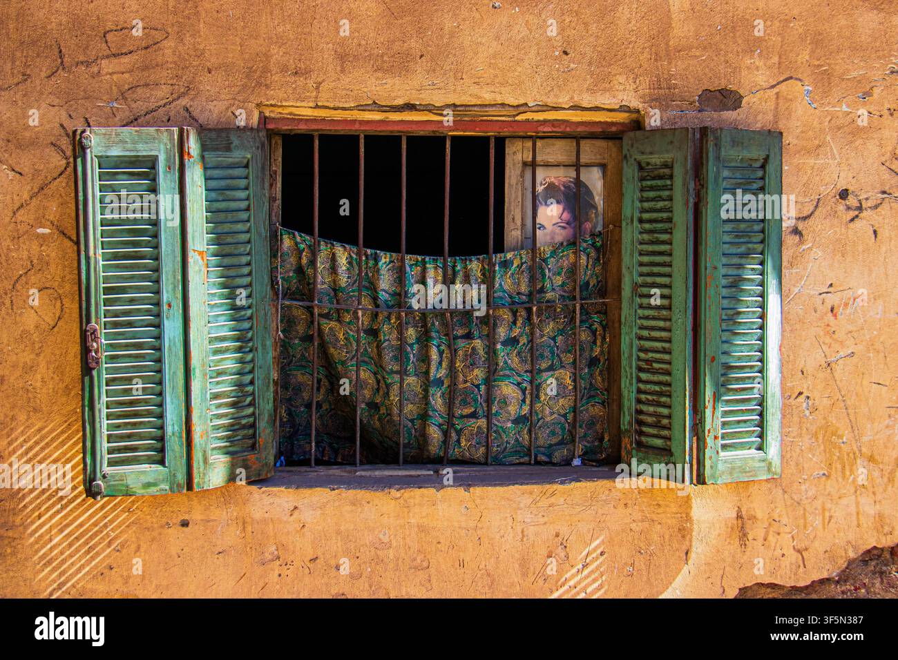Ein verwittertes Fenster mit abgeblätterten grünen Holzläden und eisernen Sicherheitsgittern in Luxor, Ägypten, fotografiert am 16. Februar 2024. Ein gemusterter Vorhang pa Stockfoto