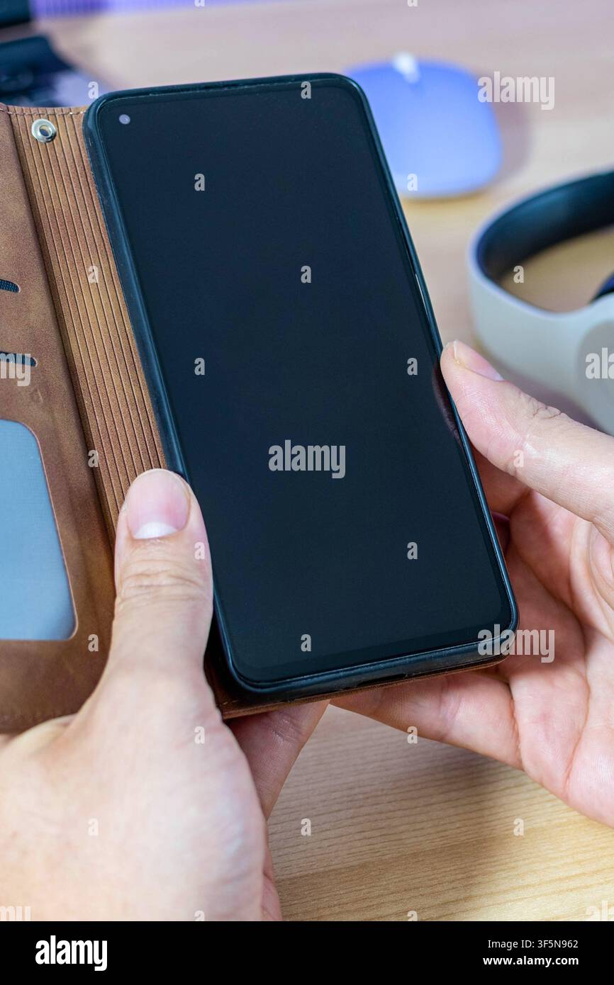 Vertikale Aufnahme von Händen, die ein Smartphone mit schwarzem Bildschirm für ein Mockup halten. Braunes Lederetui auf einem Bürotisch mit Kopfhörern. Stockfoto