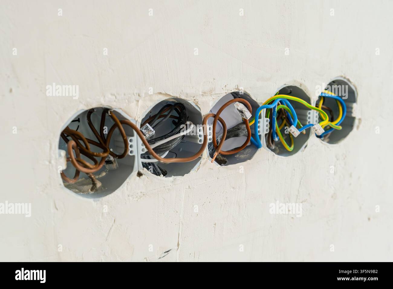Elektrikleitungen, die in mehreren Klemmpaneln angeordnet sind, die in eine Wand eingebettet sind. Freigelegte Kabel enthalten Farben für Strom-, Neutral- und Erdungsleitungen, die die Vorbereitung für die elektrische Installation anzeigen. Stockfoto