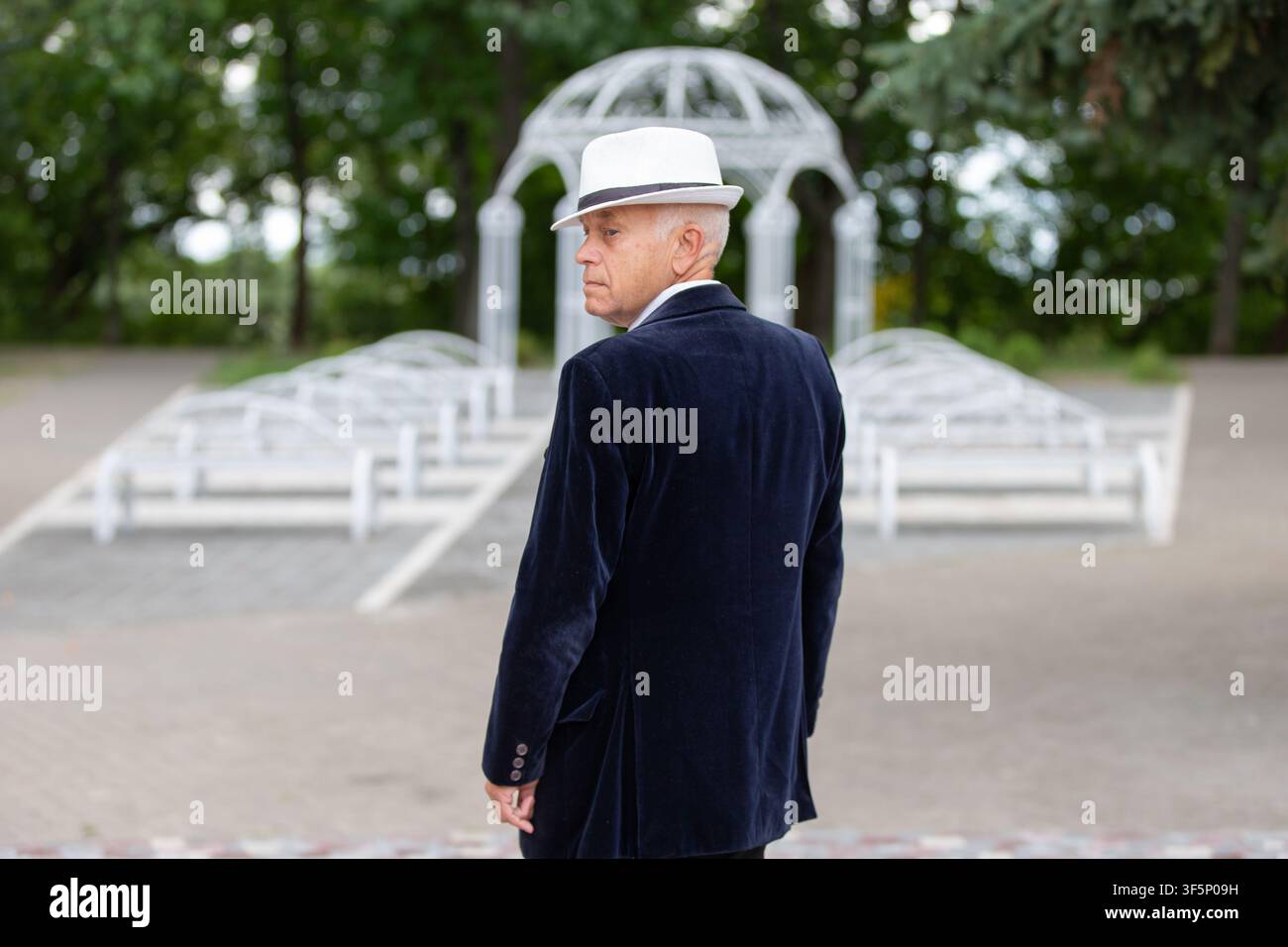 Ein älterer Mann in einem dunklen Samtanzug und einem weißen Hut im Park, der über seine Schulter blickt. Diese elegante Gestalt steht im Kontrast zur natürlichen Umgebung Stockfoto