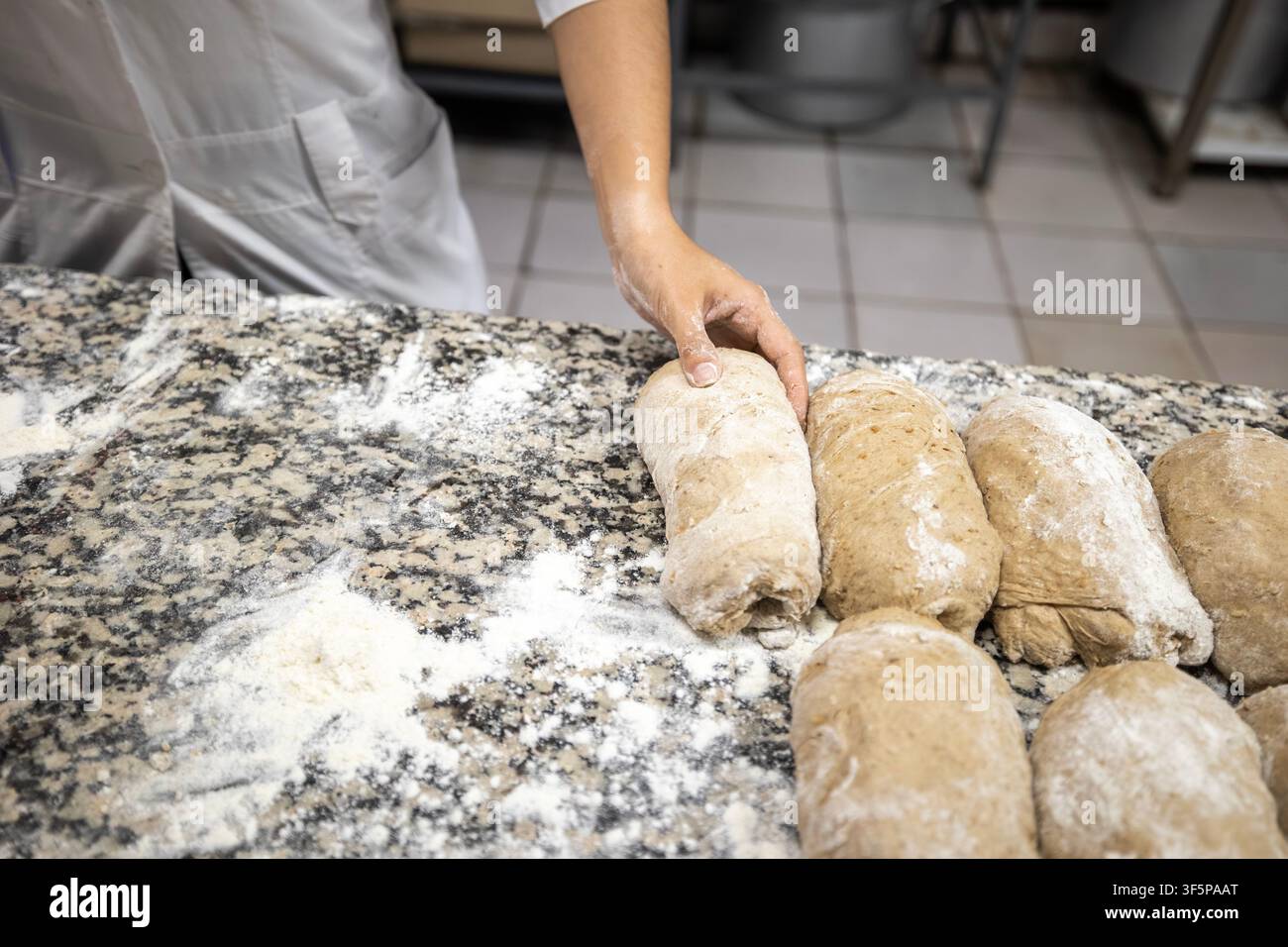 Professioneller Bäcker knetet frische Teigbrote auf einer Granitküchenoberfläche in einer gewerblichen Nahrungsmittelzubereitungsanlage mit weißer Küchenuniform und Stockfoto