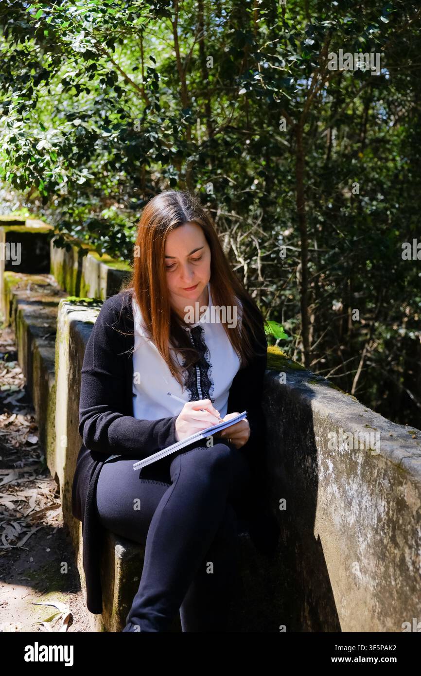 Frau schreibt Notizen in ein Notizbuch, während sie im Wald im Freien sitzt Stockfoto