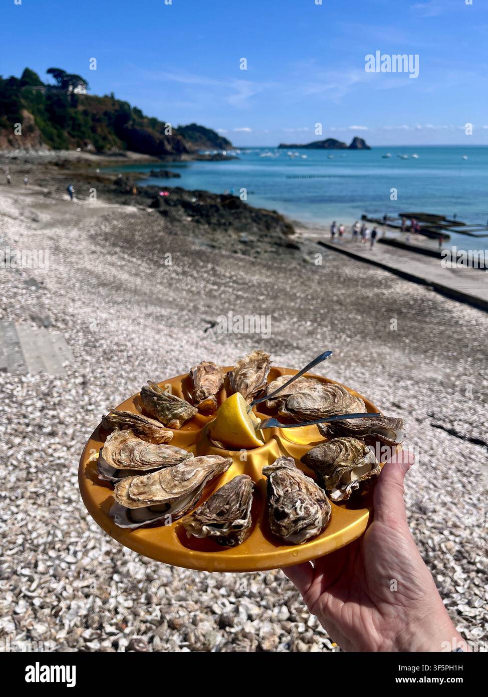 Hält einen runden Teller mit einem Dutzend frischer Austern von den Ständen im Hafen von Cancale in Frankreich mit den Austernbanken von Cancale im Hintergrund - Smartphone-aufgenommenes Stockfoto