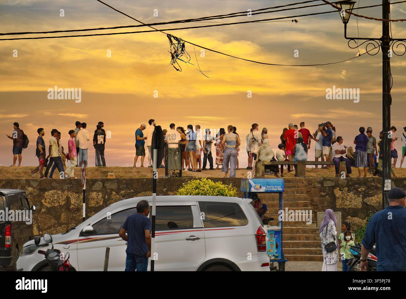 Galle Fort, Sri Lanka – Menschen stehen auf dem Wall in der Rampart Street und beobachten den Sonnenuntergang über dem Indischen Ozean, mit geparkten Autos im Vordergrund Stockfoto