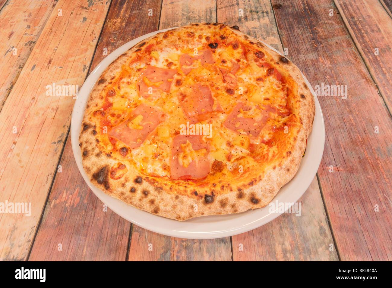 Hawaiianische Pizza mit Schinken und Ananas auf einem weißen Teller über einem rustikalen Holztisch. Stockfoto