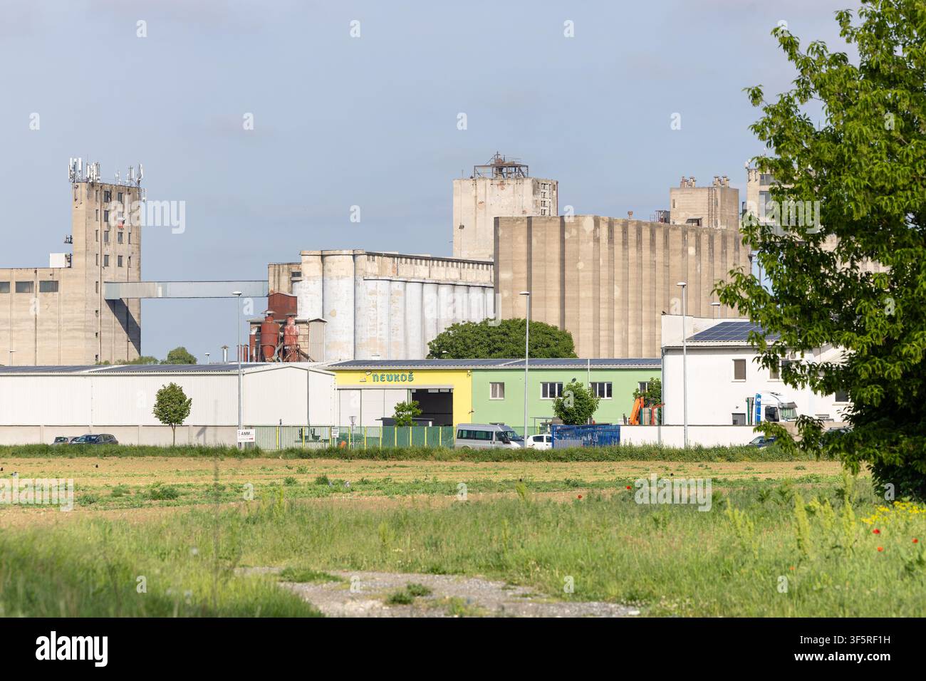 Vinkovci, Kroatien - 08. Mai 2026: Eine Reihe von Beton-Silos und Industrie-Lagerhallen ragt hinter einem Grasfeld auf, mit Lieferwagen, Beladung sind Stockfoto