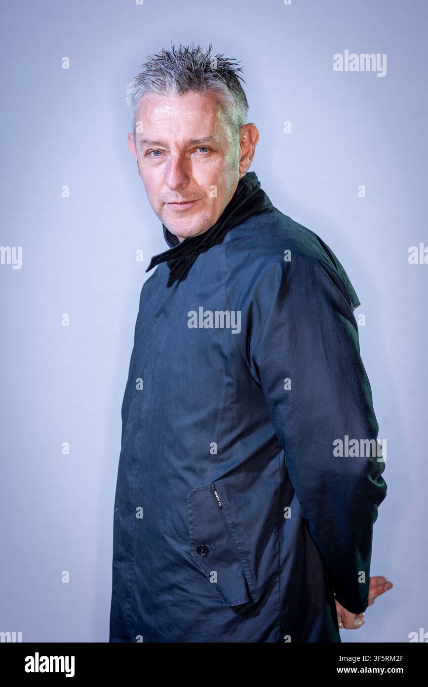 Stuart Kelly steht seitlich in einer Studioeinstellung und trägt eine schwarze Jacke. Er schaut selbstbewusst in die Kamera und zeigt einen ernsten Ausdruck. Der Hintergrund Stockfoto