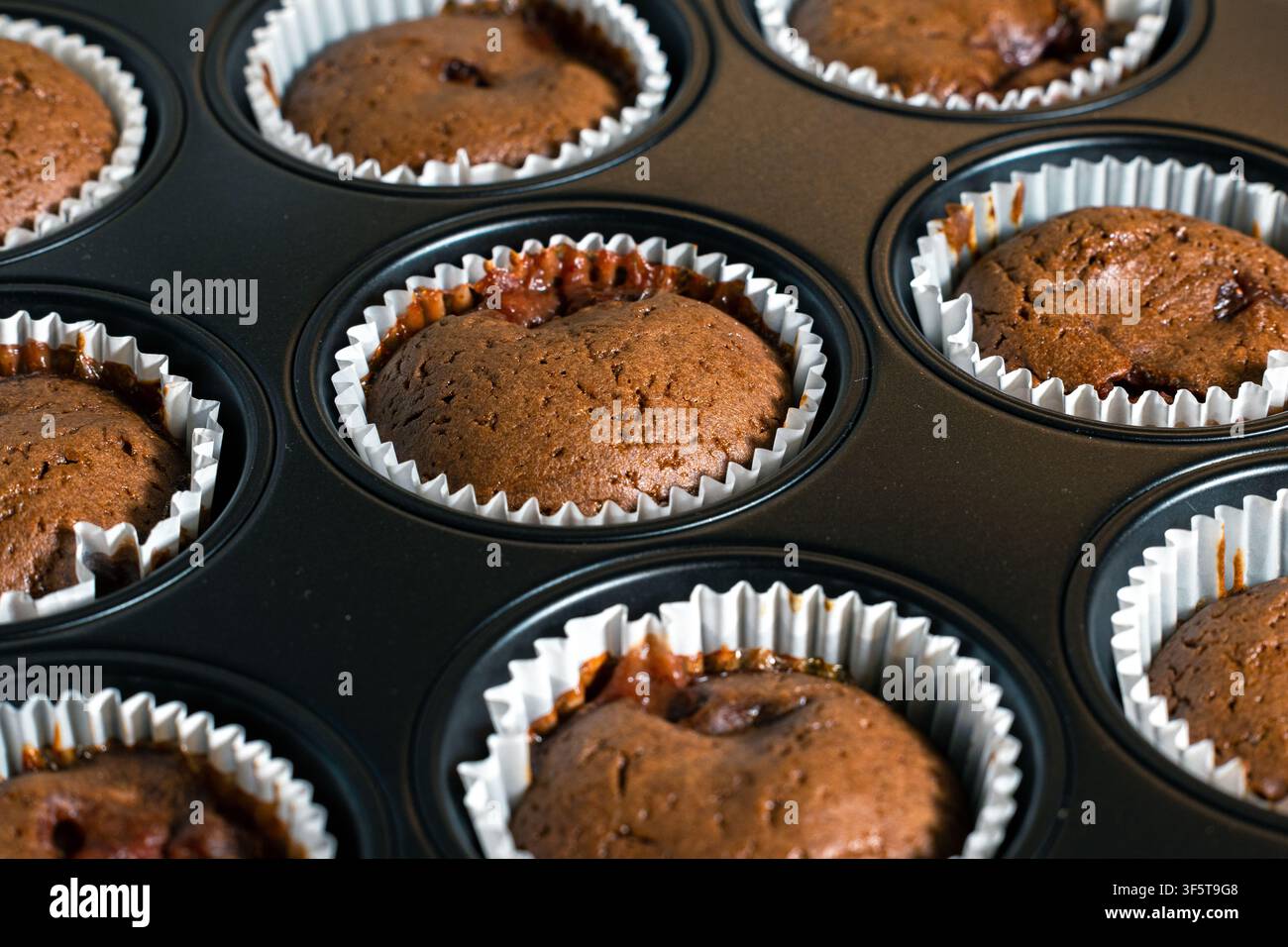 Schokoladen-Muffins ruhen in einem dunklen Tablett mit weißen Papierförmchen. Braune Oberseiten zeigen eine rissige Textur und kleine Bereiche mit geschmolzener Füllung. Stockfoto