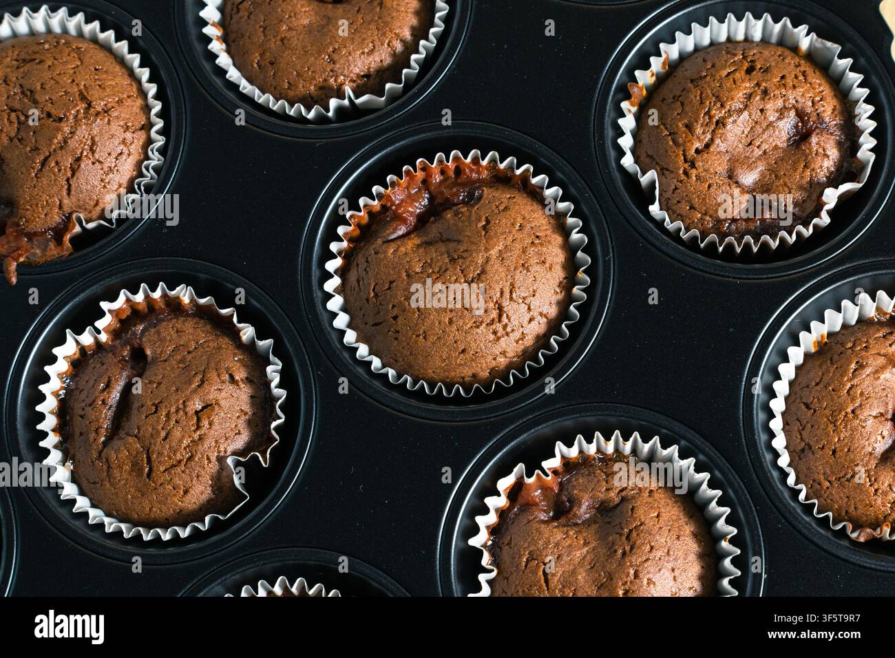 Heiße Schokoladen-Muffins backen in einem dunklen Blech mit weißen Papierförmchen. Braune Oberseiten zeigen eine rissige Textur und kleine Bereiche mit geschmolzener Füllung. Stockfoto