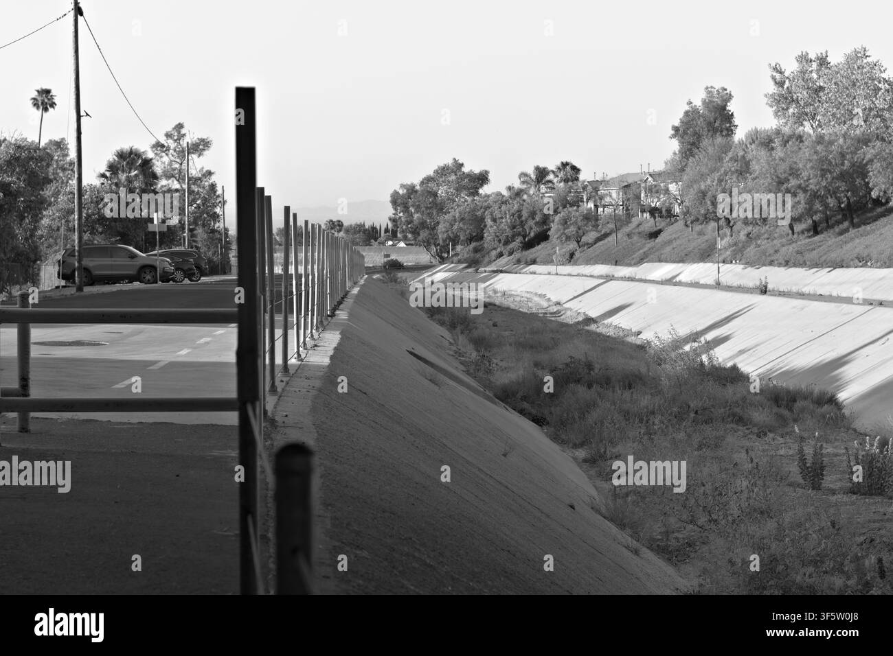Ein trockener Beton-Überwasserkanal verläuft neben einem Parkplatz und einem landschaftlich gestalteten Hügel, gesäumt von Barrieren, Unkraut, Bäumen und nahe gelegenen Häusern. Stockfoto