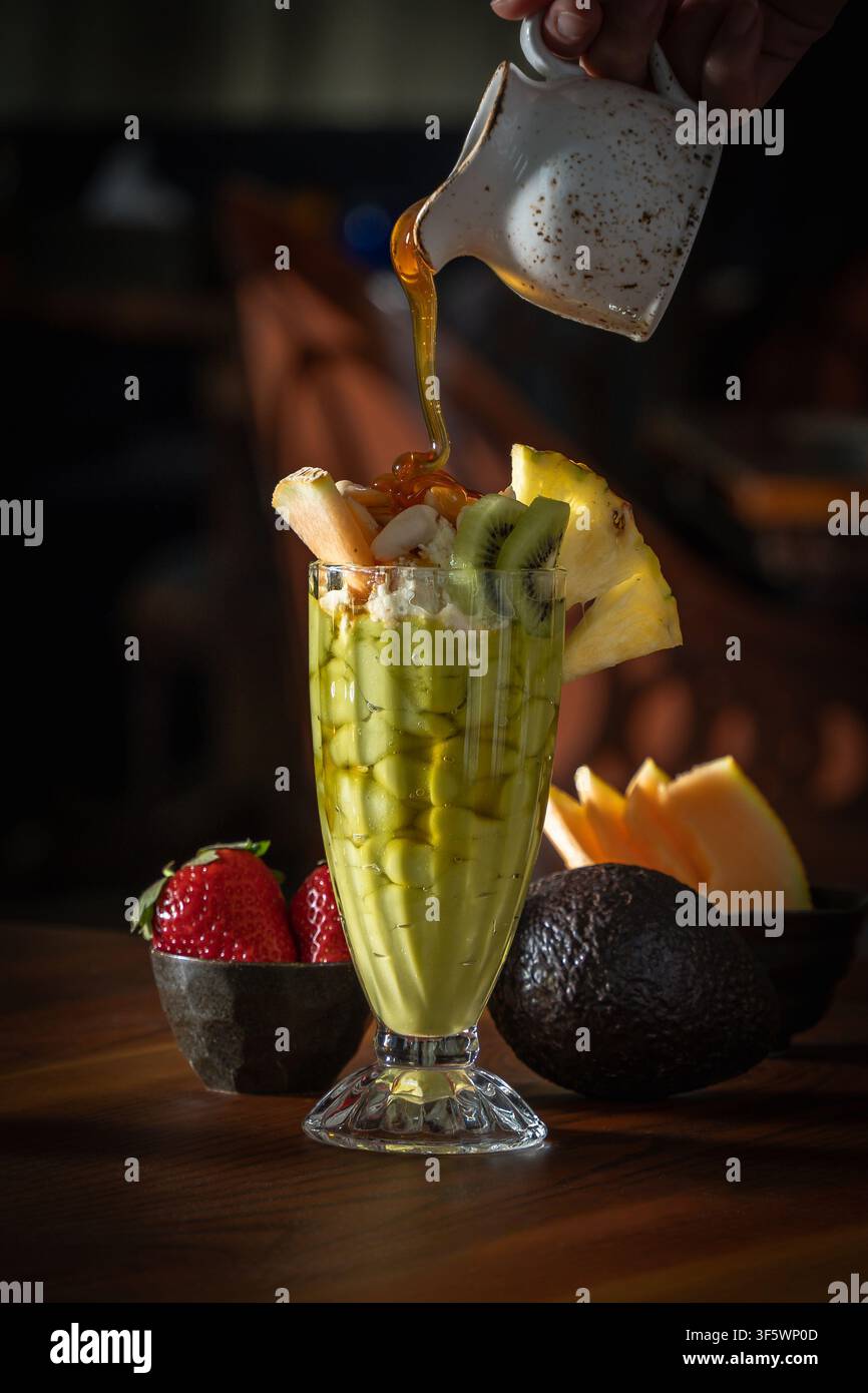 Honig, der aus einem Keramikkanister über einen Avocado-Cocktail geträufelt wird, der mit Sahne, Mandeln, Kiwi und Ananas gekrönt ist, mit frischer Avocado, Melone und Erdbeeren Stockfoto