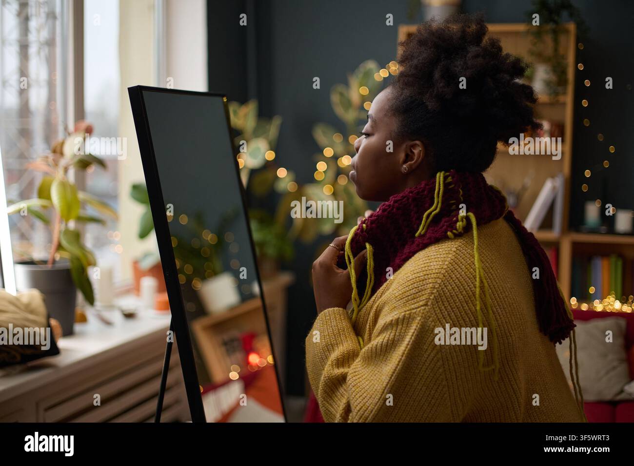 Junge afroamerikanische Frau schaut in den Spiegel, während sie einen gestrickten Schal anprobiert, und bereitet sich darauf vor, auszugehen. Bild repräsentiert Selbstfürsorge und Wintermode, ideal für Einzelhandelsmarketing Stockfoto