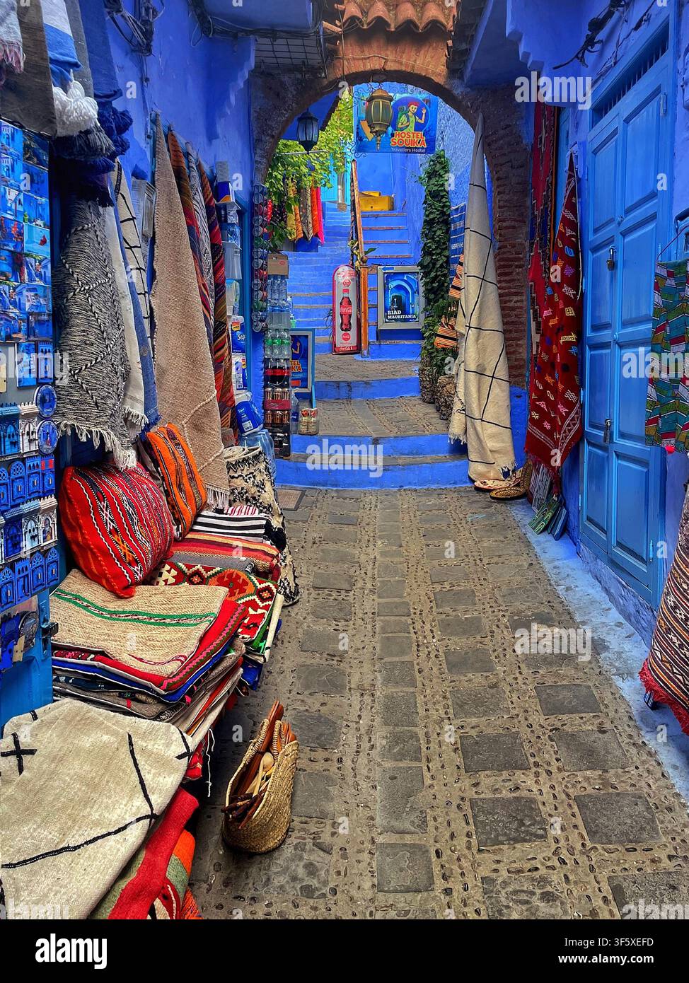 Bunte Textilien und abgefüllte Waren, die entlang einer lebendigen blauen Gasse in Chefchaouen, Marokko, ausgestellt sind. 4. August 2025. Stockfoto
