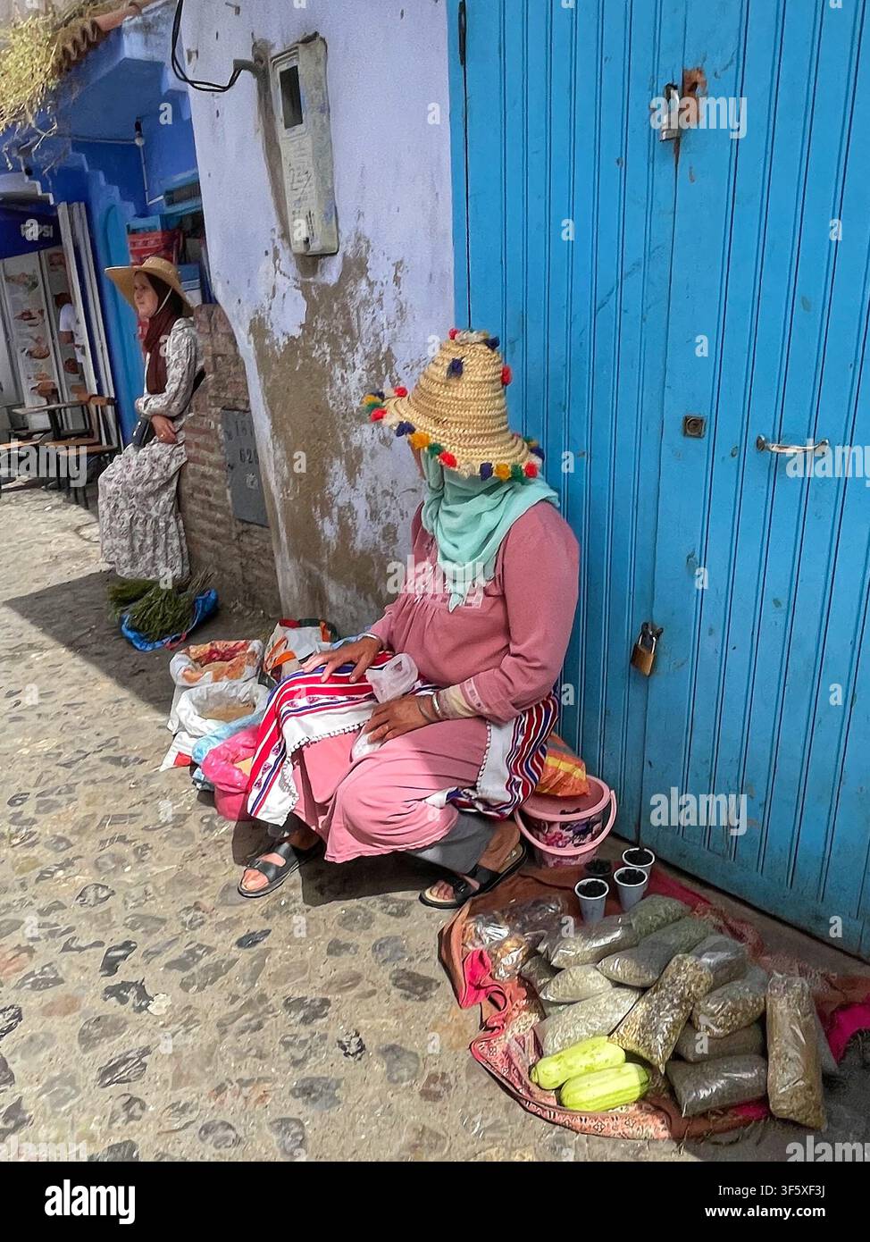 Einheimische Frau verkauft Kräuter und Gewürze auf einer Kopfsteinpflasterstraße in Chefchaouen, Marokko, mit traditionellem Hut, 4. Aug. 2025 Stockfoto