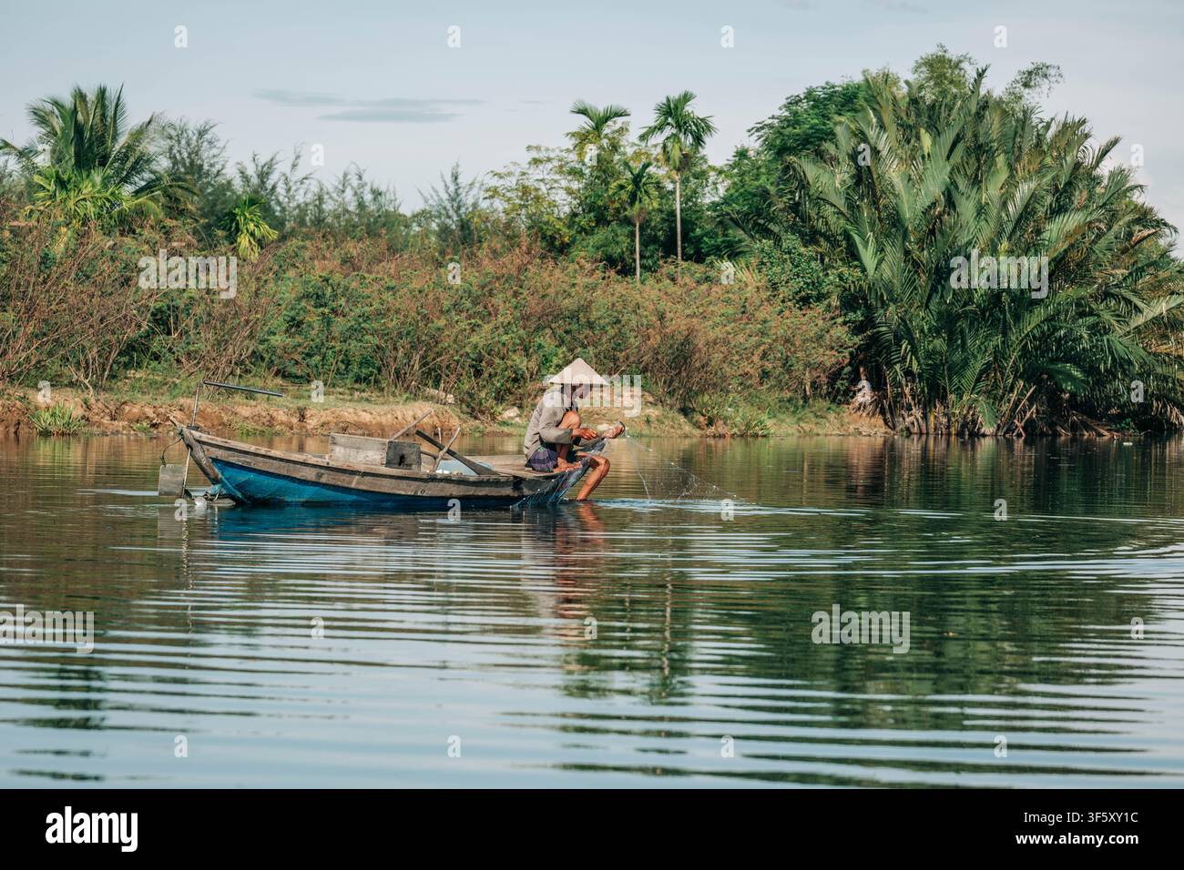 Hoi An, Vietnam, 26. Juli 2026. Ein Mann mit traditionellem Kegelhut sitzt am Rand seines blauen Bootes und pflegt ein Fischernetz in einer weiten, Stockfoto