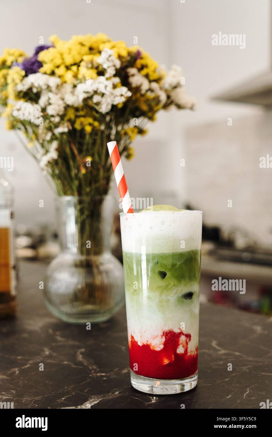 Schichtweiser Erdbeer-Matcha-Latte mit cremiger Sahnehaube, serviert mit einem gestreiften Papierstrohhalm. Erfrischendes Spezialkaffegetränk im Nahaufnahme. Stockfoto
