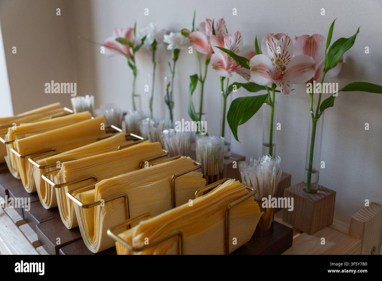 Gemütliche Cafékasse mit gelben Servietten in Messinghaltern und rosa Alstroemeria-Blumen in Glasvasen auf Holzpräsentation mit sanftem Licht. Stockfoto