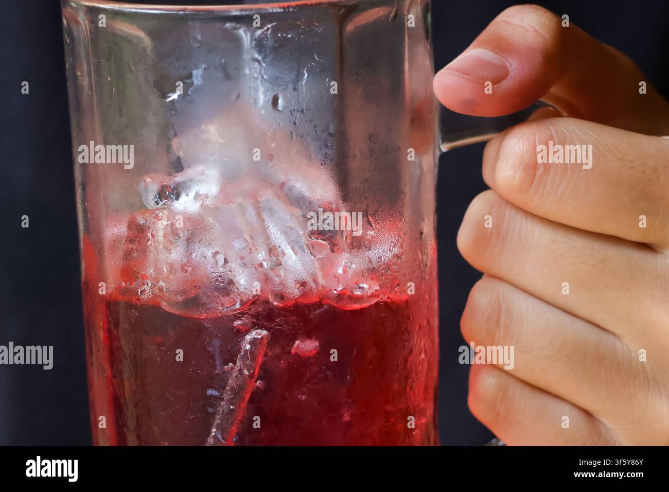 Ein gläserner Becher gefüllt mit rotem Eiskaltgetränk mit schmelzendem Eis und Hand, die den Griff hält, erfrischendes kaltes Getränk mit Kondenswasser und heller Farbe Stockfoto