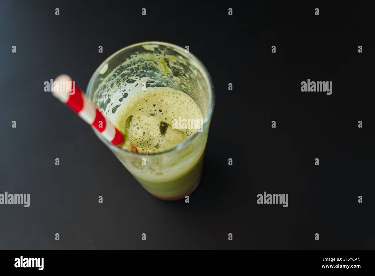 Halb fertiger Eismatcha-Latte mit Eiswürfeln und einem gestreiften Papirostrohhalm in einem klaren Glas. Erfrischendes grünes Teegetränk, serviert in einem modernen Café. Stockfoto