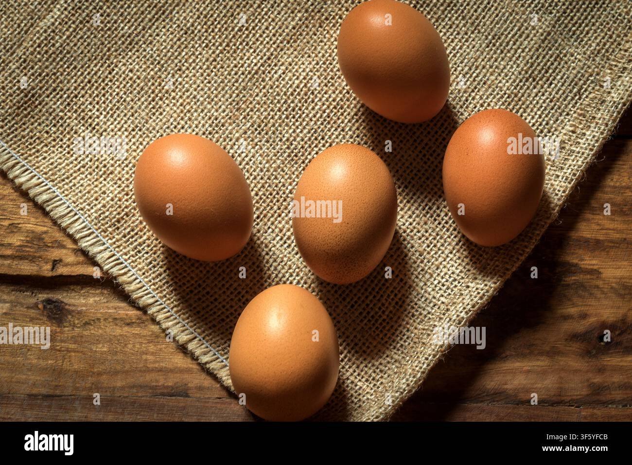 Fünf perfekte braune Eier auf einem Hintergrund aus Leinen und Holz; die Texturen werden in klaren Details dargestellt und durch den Einsatz von Licht und Schatten betont. Stockfoto