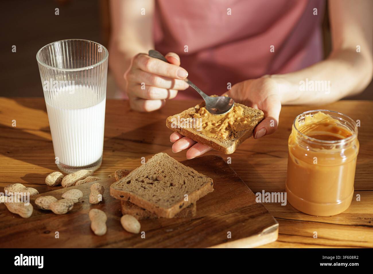 Nahaufnahme einer Frau, die knusprige Erdnussbutter auf getoastetem Brot streicht, mit Milch und Erdnüssen auf dem Tisch, um ein einfaches hausgemachtes Frühstück oder einen Snack zu veranschaulichen Stockfoto