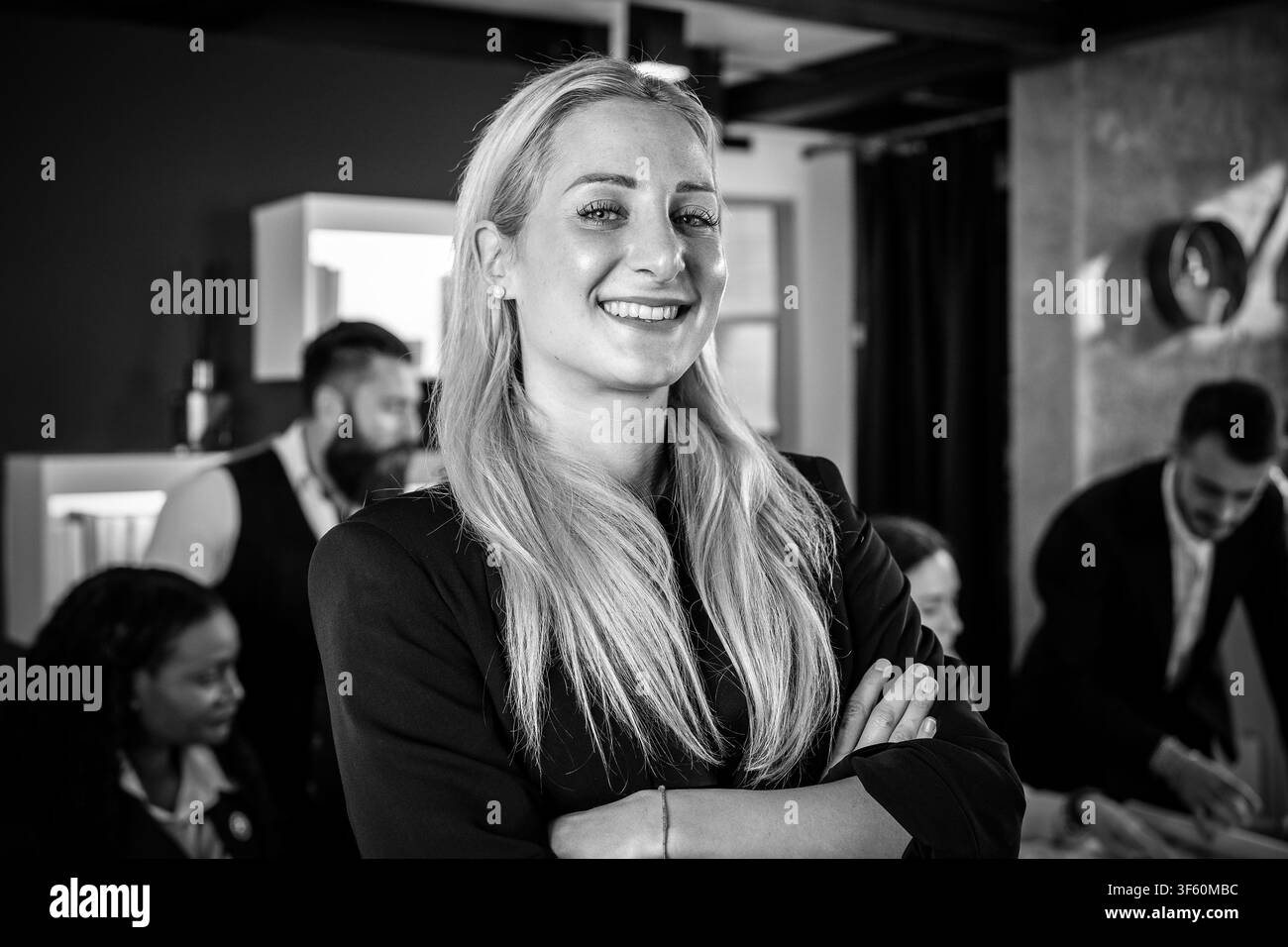 Lächelnde blonde Geschäftsfrau, die in einem modernen Büro arbeitet, mit dem multiethnischen Team hinter ihr - Konzept von Führung und Teamarbeit Ziele erreichen - Stockfoto
