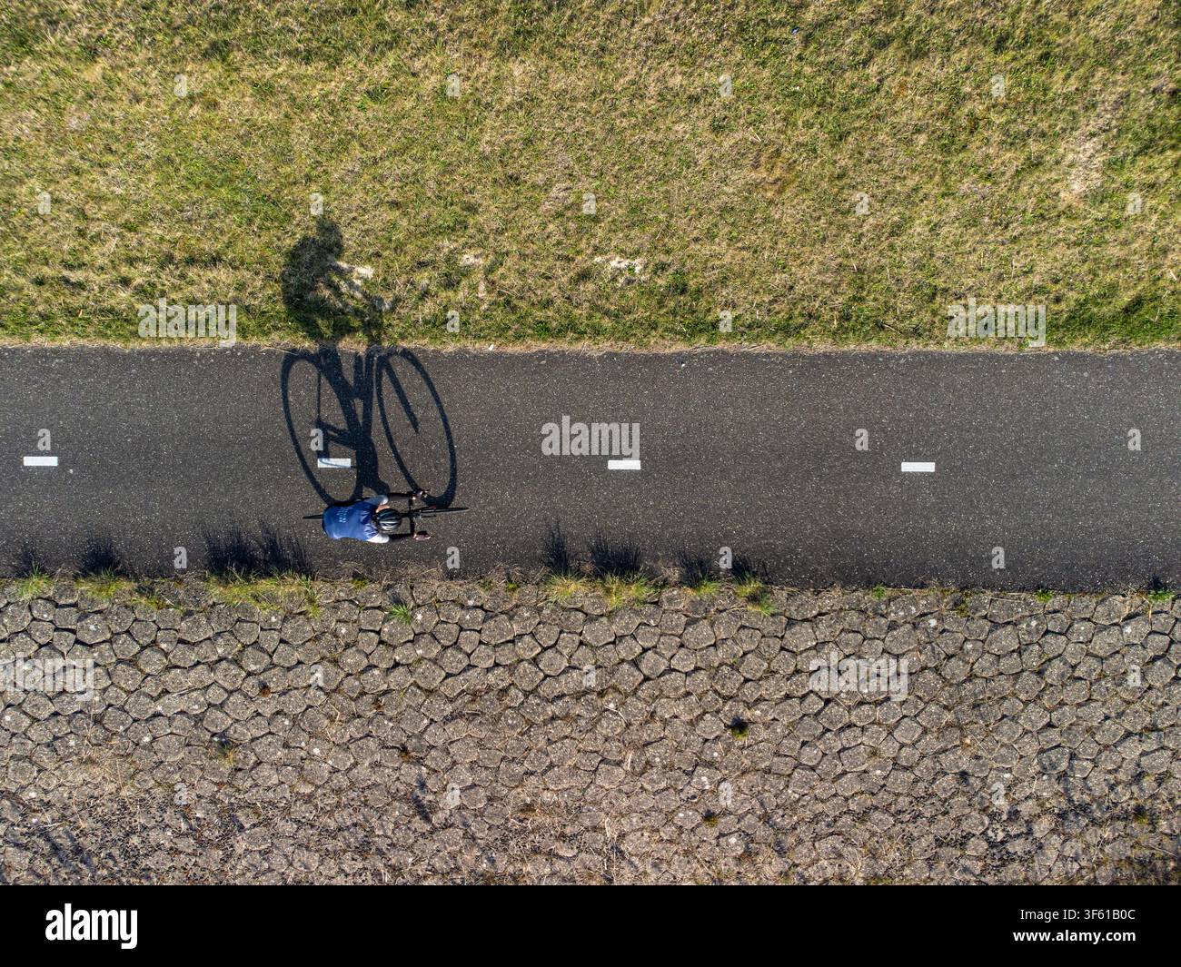 Luftaufnahme eines Radfahrers, der entlang einer asphaltierten Straße fährt, mit einem langen Schatten, der auf den Boden fällt, begrenzt von einem grasbewachsenen Straßenrand und einem stein gepflasterten Damm i Stockfoto