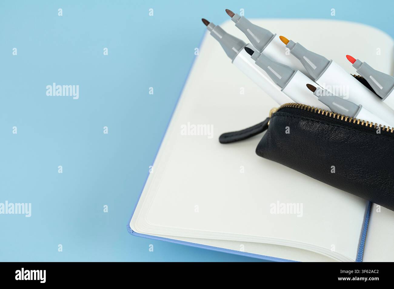 Bunte Kunstmarker in einem schwarzen Federmäppchen, die auf einem offenen Notizbuch auf einem hellblauen Hintergrund angeordnet sind Stockfoto