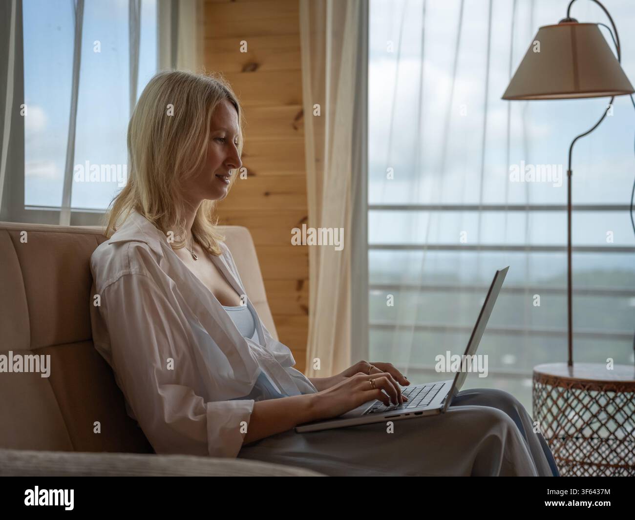 Blonde Frau arbeitet von zu Hause aus, sitzt bequem auf einer Couch und tippt auf einem modernen Laptop, genießt eine entspannte und produktive Fernarbeit oder Freelancing Stockfoto