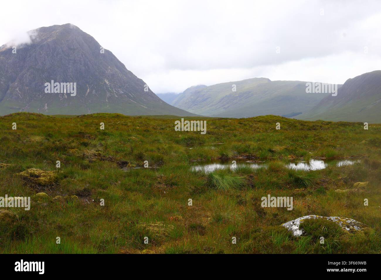 Moorland in den schottischen Highlands mit grasbewachsenem, feuchtem Gelände und einem großen felsigen Berg, der teilweise von tiefen Wolken verdeckt ist, die eine geschichtete Naturlandschaft bilden Stockfoto