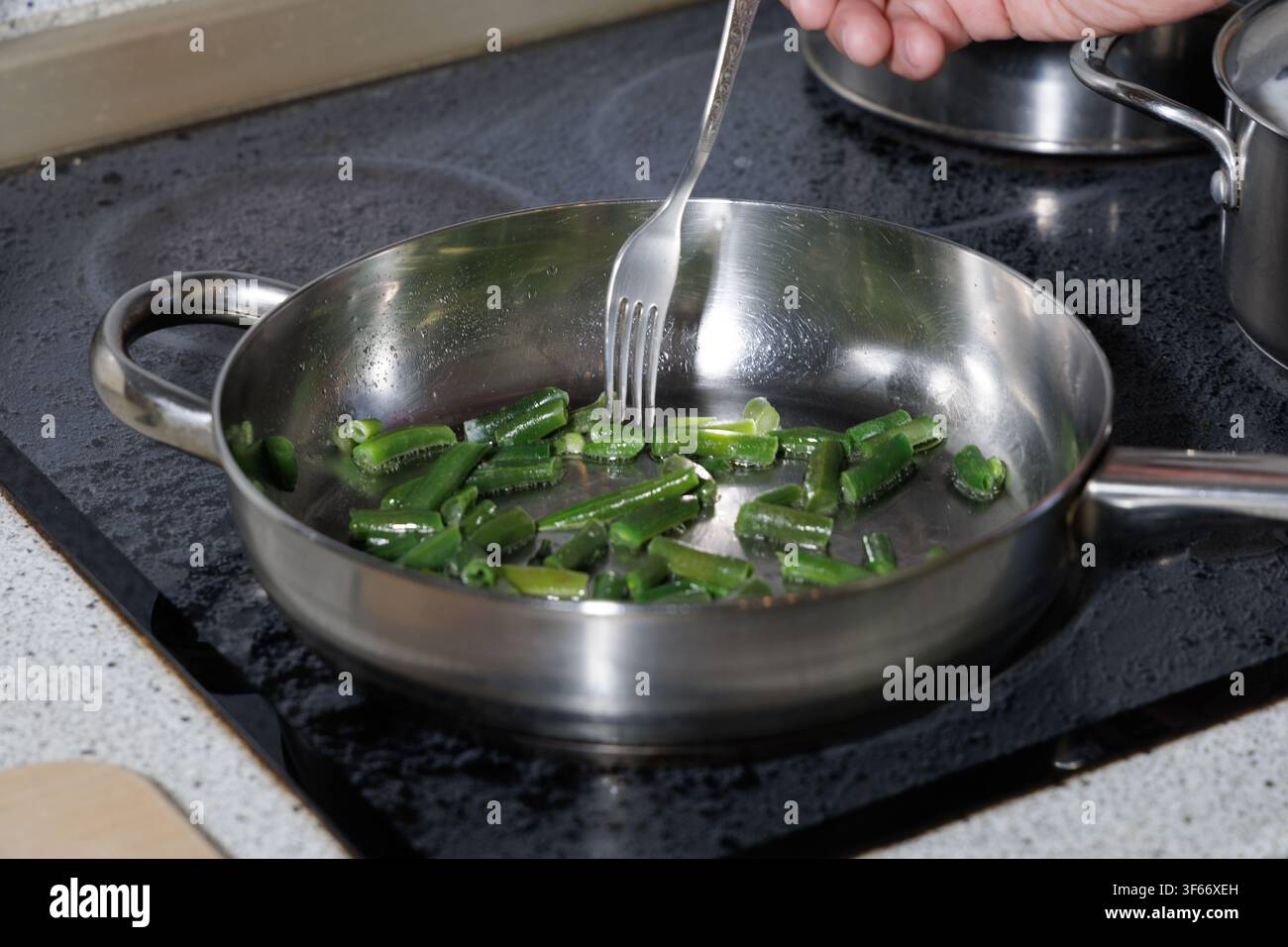 Nahaufnahme einer Person, die frische gehackte grüne Bohnen mit einer Gabel in einer Edelstahlbratpfanne auf einem modernen Küchenherd kocht. Stockfoto
