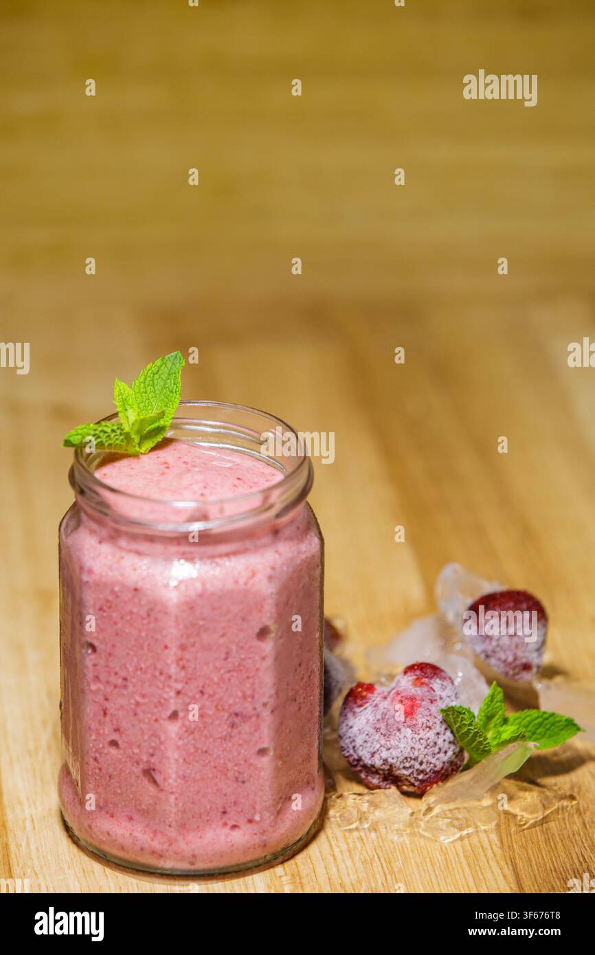 Frischer organischer Erdbeersmoothie, serviert in einem Glasbehälter, garniert mit grünen Minzblättern, platziert in der Nähe von gefrorenen Beeren und Eis auf einem Holztisch mit Oberfläche Stockfoto