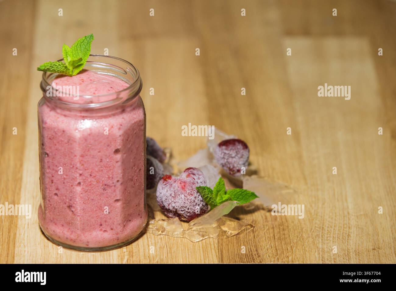 Frischer Erdbeersmoothie serviert in einem Glasbehälter, garniert mit Minzblättern, platziert in der Nähe von gefrorenen Beeren und Eis auf einem rustikalen Holztisch mit Kopierraum Stockfoto