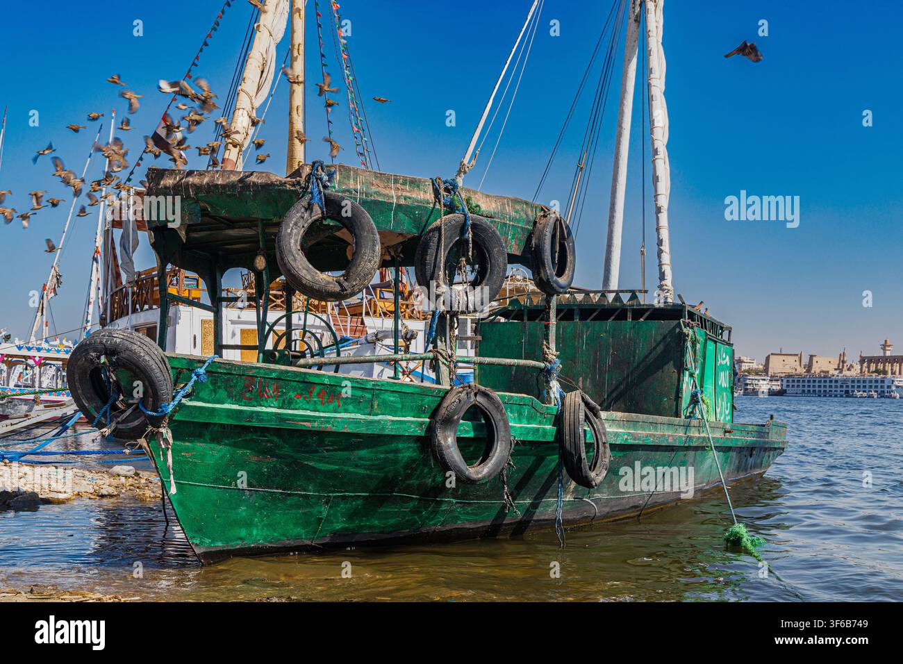 Ein verwittertes grünes Holzboot mit Reifenfendern, das am Nil in Luxor, Ägypten, festgemacht ist, mit Vögeln im Flug und dem Luxor-Tempel im dunstigen Hintergrund. Stockfoto
