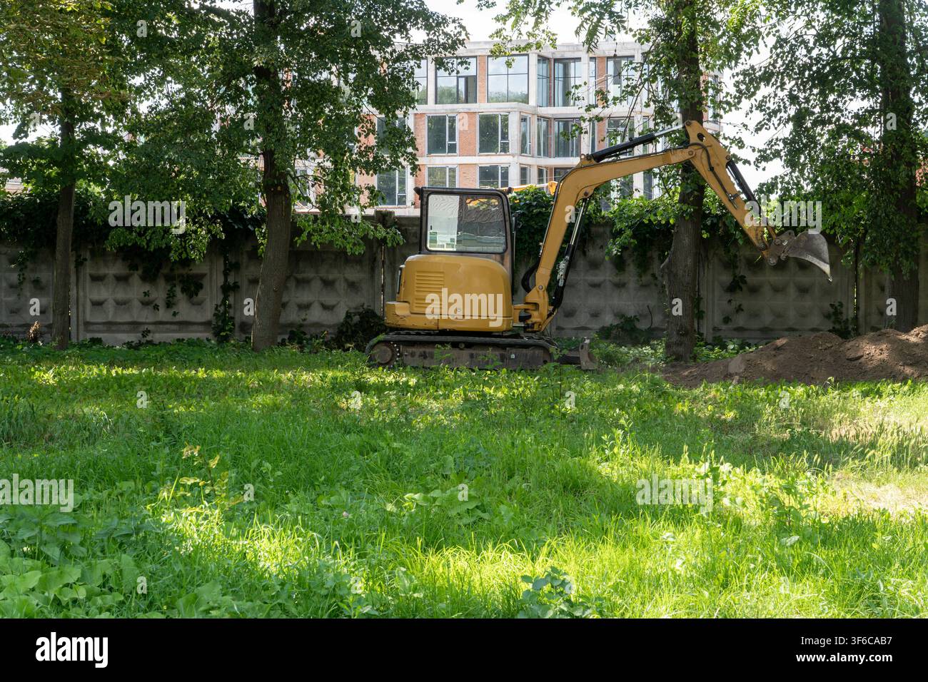 Kleiner Kettenbagger mit gehobenem Hydraulikarm gräbt Boden aus und schafft einen Erdhaufen in der Nähe von grünen Bäumen an der Baustelle. Stockfoto