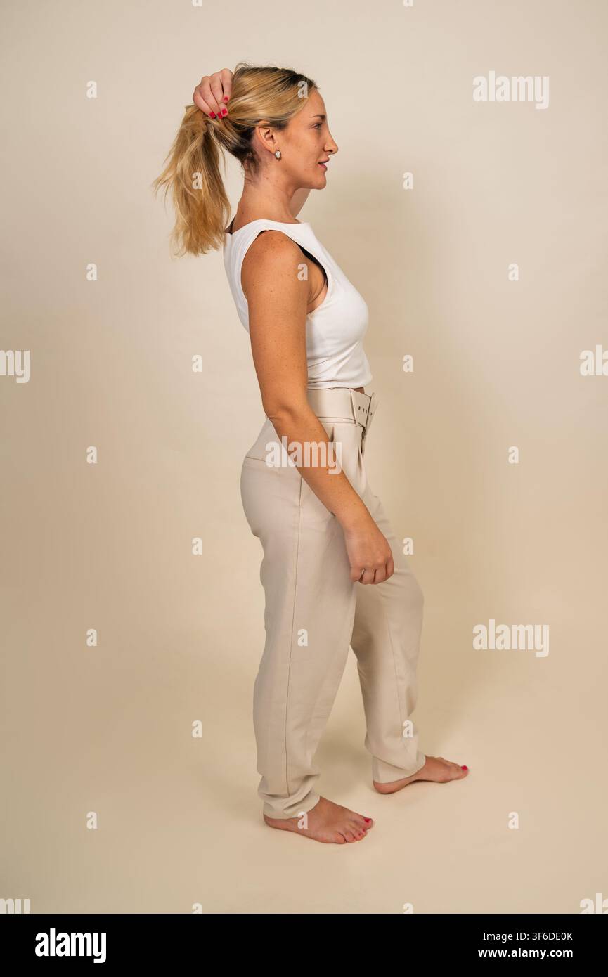 Frau, die ihr blondes Haar zu einem hohen Pferdeschwanz bindet, vor einem schlichten Hintergrund Stockfoto