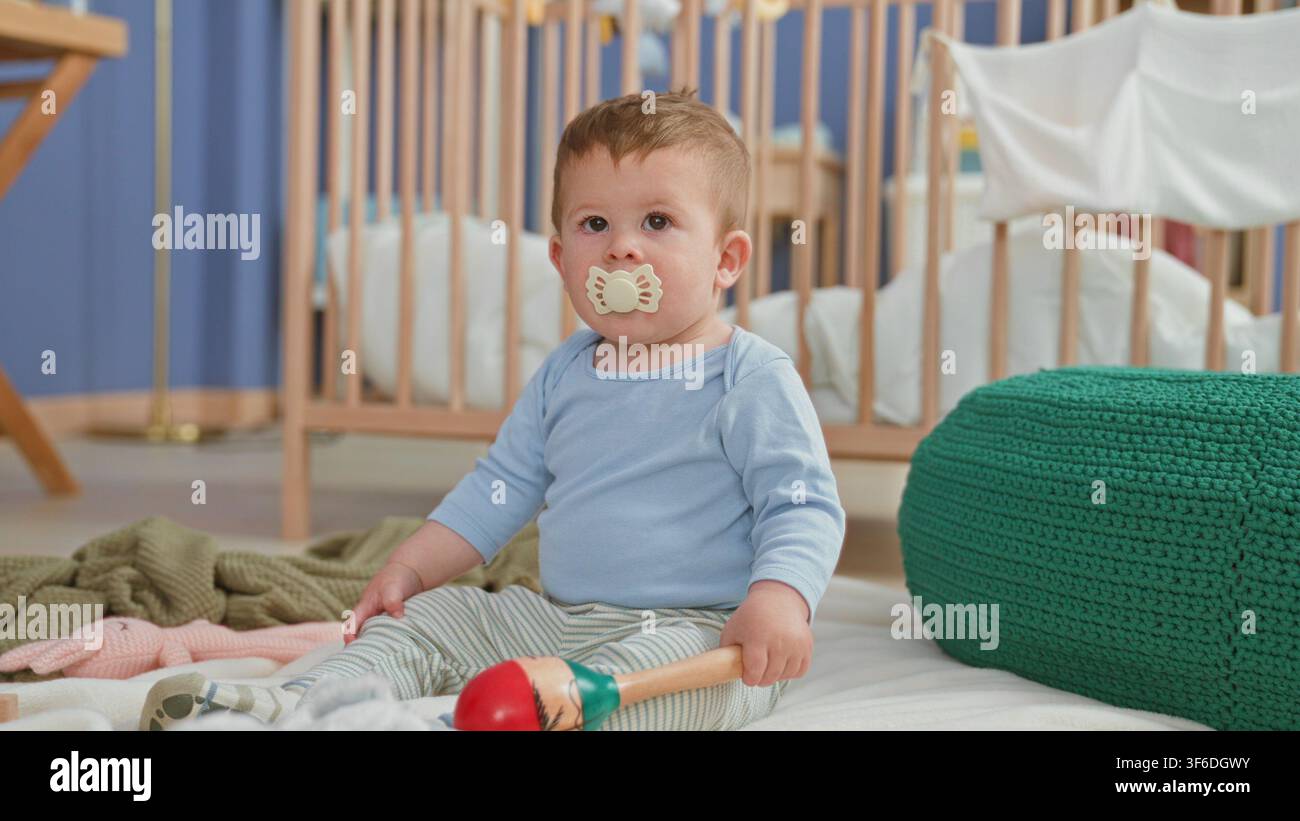 Kleiner Junge sitzt mit Schnuller und hält eine Maracas, streckt die Hand in Richtung Bett in einem Gebäude; unschuldige Neugier. Stockfoto