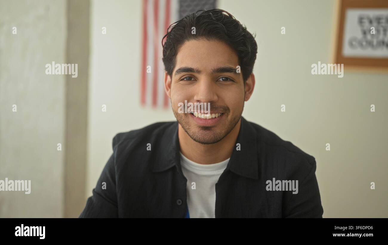 Mann lächelt, zeigt die Zähne und schaut in die Kamera neben einer amerikanischen Flagge und einem „Jede Stimme zählt“-Plakat in einem Wahllokal; bürgerstoleranter Stolz. Stockfoto