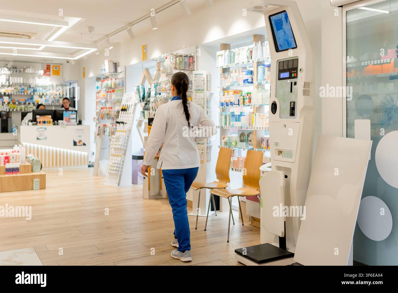 Eine weibliche Apothekenmitarbeiterin geht durch eine helle moderne Apotheke, vorbei an Medikamentenregalen, Kundenbestuhlung und einem automatisierten Gesundheitsservice-Kiosk. Stockfoto